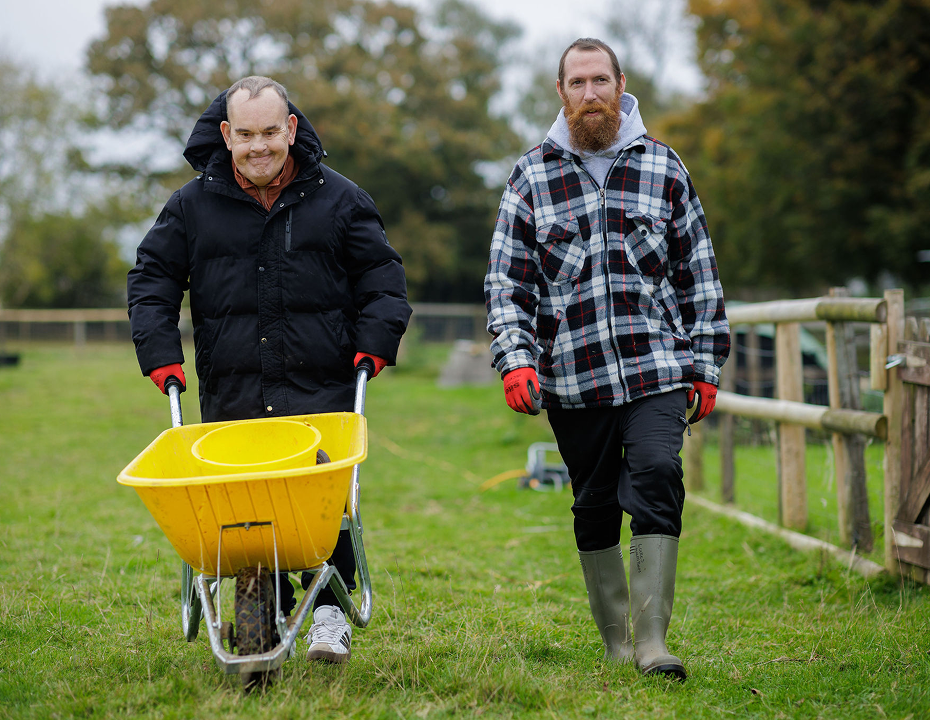 Two people outdoors in a grassy field, one pushing a yellow wheelbarrow, both wearing coats and gloves.
