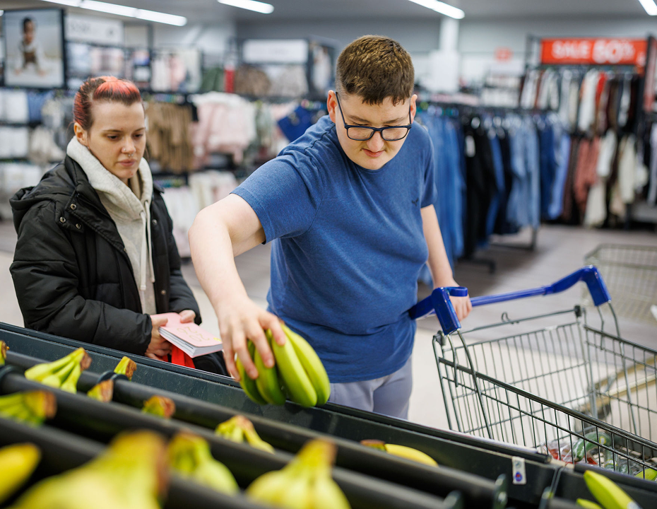Person selecting bananas in a supermarket while pushing a shopping trolley, with another person standing nearby. Supported living care services in Caerphilly.